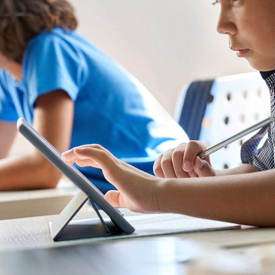 Close up cut shot of concentrated diligent elementary schoolboy touching using tablet device sitting in classroom with group of schoolchildren.